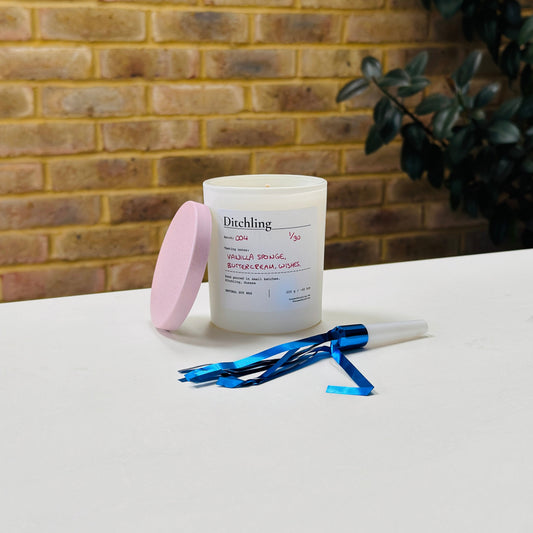 Ditchling Batch 004 candle, 1/30, “Vanilla Sponge, Buttercream, Wishes”, hand poured in small batches in Ditchling, Sussex, with natural soy wax, pink sparkle lid and white glass jar, photographed on a white table against a warm brick wall with a leafy plant in the background.