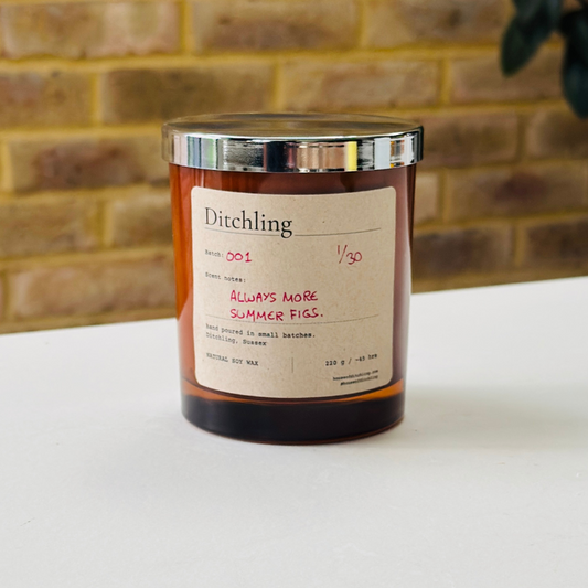 Ditchling “Always More Summer Figs” candle, Batch 001, 1/30, in an amber glass jar with silver lid. Label reads “Scent notes: ALWAYS MORE SUMMER FIGS.”, “hand poured in small batches. Ditchling, Sussex”, “NATURAL SOY WAX”, “220 g / ~45 hrs”, “houseofditchling.com” and “@houseofditchling”. Photographed on a white surface against a warm brick wall with greenery.