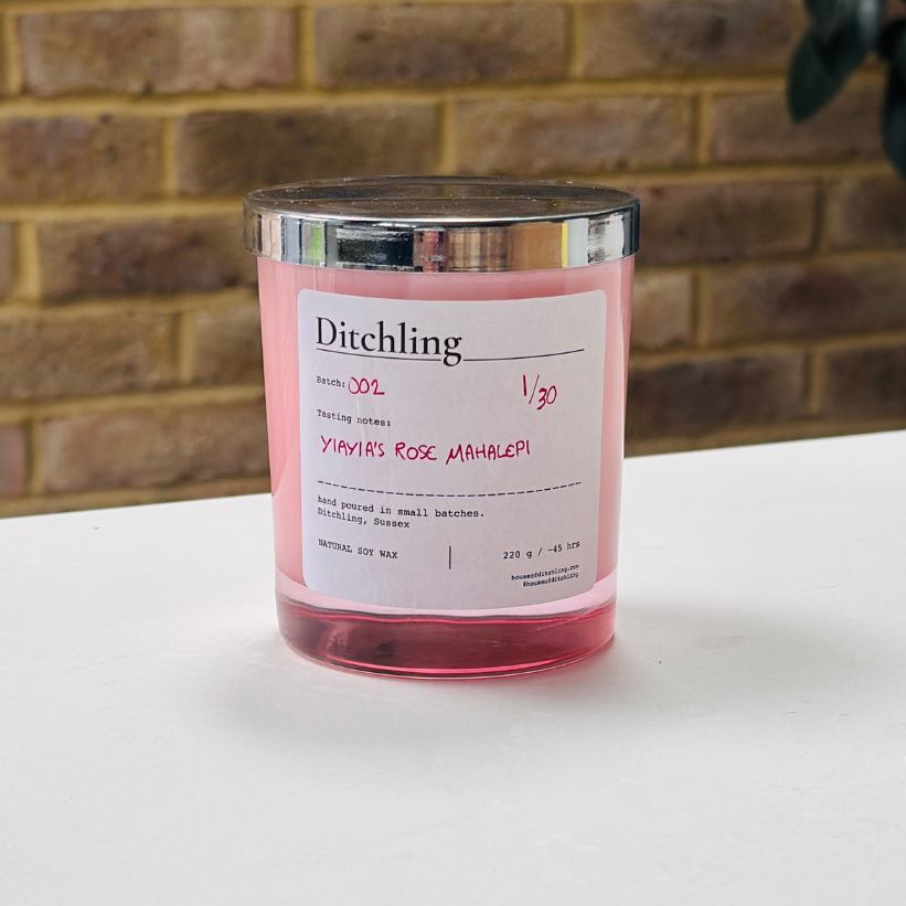 Ditchling “Yiayia’s Rose Mahalepi” candle, Batch 002, 1/30, in a pink glass jar with silver lid. Label reads “Tasting notes: YIAYIA’S ROSE MAHALEPI”, “hand poured in small batches. Ditchling, Sussex”, “NATURAL SOY WAX”, “220 g / ~45 hrs”, “houseofditchling.com” and “@houseofditchling”. Photographed on a white surface against a warm brick wall with greenery.