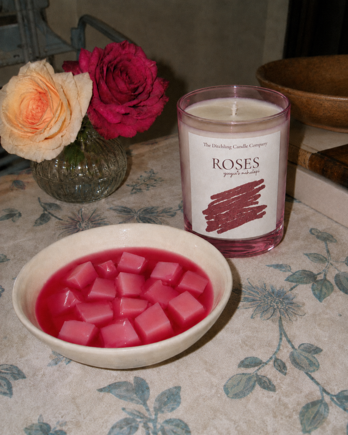 The Ditchling Candle Company ROSES candle beside a bowl of pink mahalepi Cypriot rose pudding, with fresh roses in a glass vase on a vintage floral tablecloth.