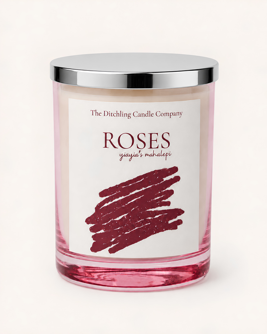 A glass jar soy scented candle with a silver lid and a white label reading 'The Ditchling Candle Company ROSES yiayia's mahalepi' featuring a burgundy scribble graphic on a light rose pink candle inside.