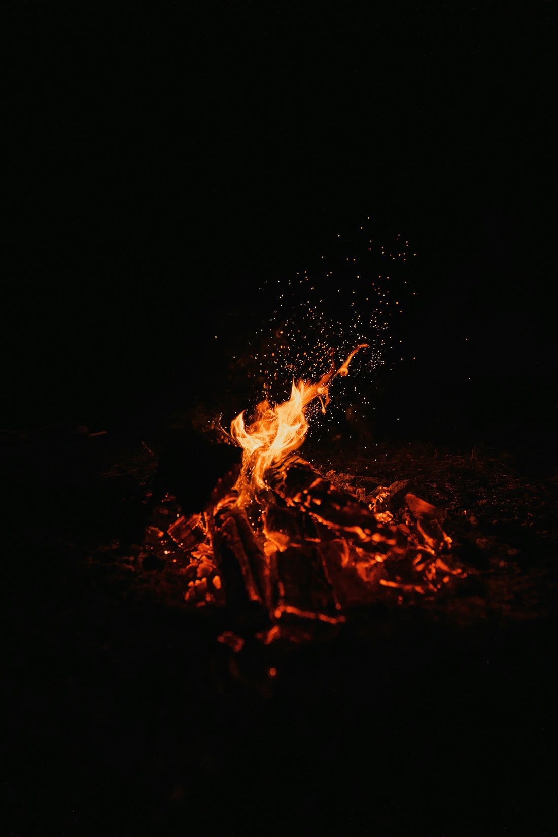 A small campfire burning in the darkness, with glowing embers, charred logs, bright orange flames and sparks rising into the night.