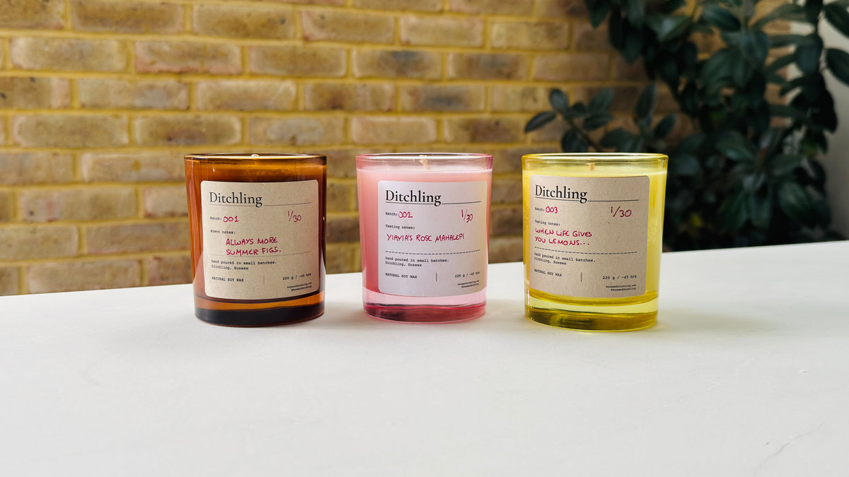 Three Ditchling brand candles in amber, pink, and yellow glass jars with white labels, arranged on a white surface against a brick wall background.