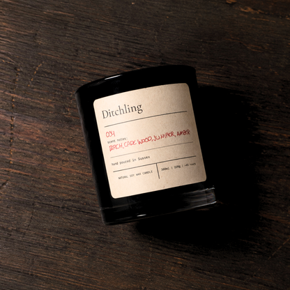 Ditchling 004 Embers scented soy candle in a black glass jar with a kraft paper label, featuring birch, cade wood, juniper and amber scent notes. Wooden background.