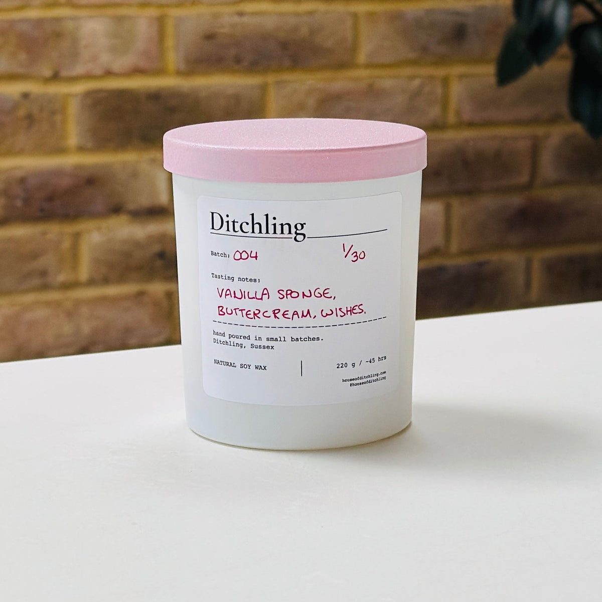 Ditchling Batch 004 candle, 1/30, “Vanilla Sponge, Buttercream, Wishes”, hand poured in small batches in Ditchling, Sussex, with natural soy wax, pink sparkle lid and white glass jar, photographed on a white table against a warm brick wall with a leafy plant in the background.