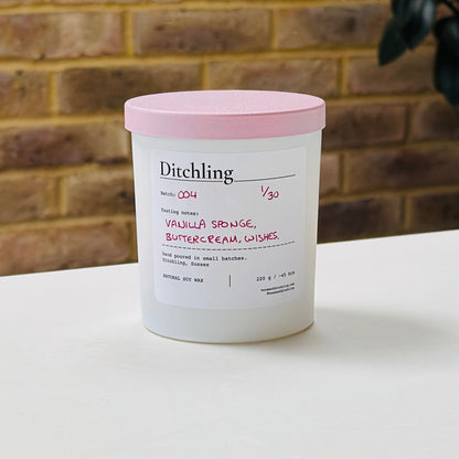 Ditchling Batch 004 candle, 1/30, “Vanilla Sponge, Buttercream, Wishes”, hand poured in small batches in Ditchling, Sussex, with natural soy wax, pink sparkle lid and white glass jar, photographed on a white table against a warm brick wall with a leafy plant in the background.