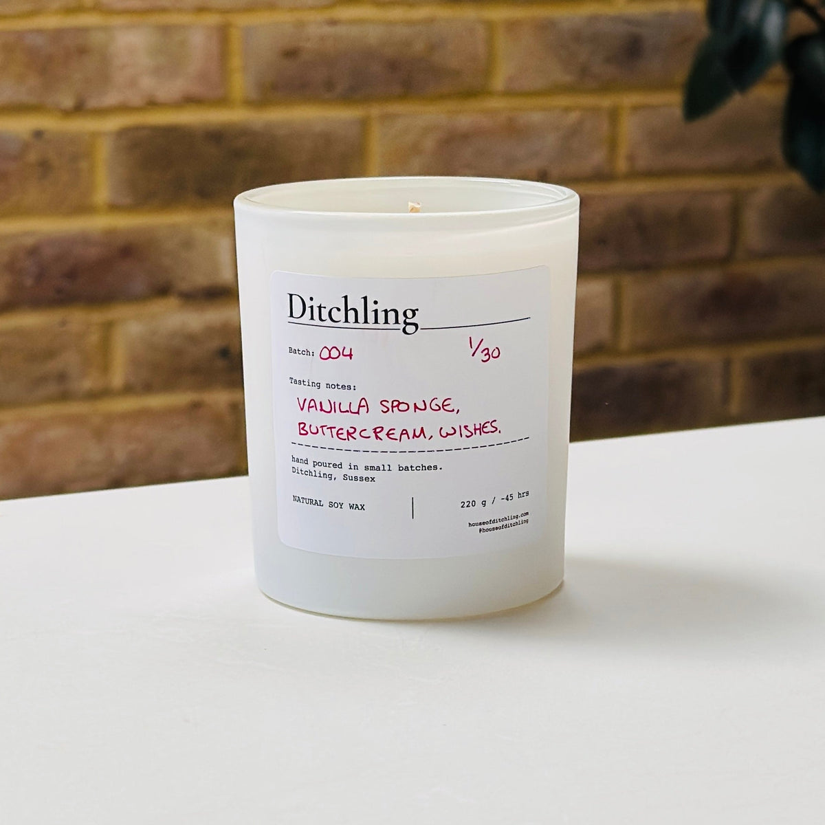Ditchling Batch 004 candle, 1/30, “Vanilla Sponge, Buttercream, Wishes”, hand poured in small batches in Ditchling, Sussex, with natural soy wax, pink sparkle lid and white glass jar, photographed on a white table against a warm brick wall with a leafy plant in the background.