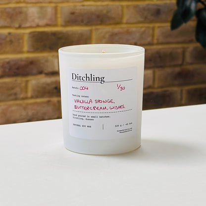 Ditchling Batch 004 candle, 1/30, “Vanilla Sponge, Buttercream, Wishes”, hand poured in small batches in Ditchling, Sussex, with natural soy wax, pink sparkle lid and white glass jar, photographed on a white table against a warm brick wall with a leafy plant in the background.