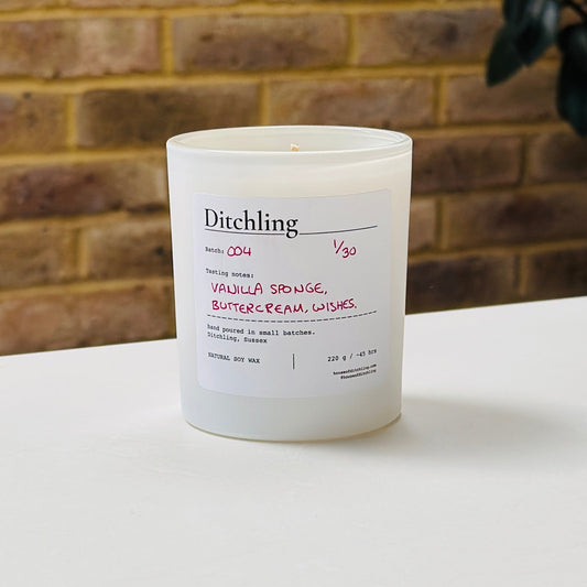 Ditchling Batch 004 candle, 1/30, “Vanilla Sponge, Buttercream, Wishes”, hand poured in small batches in Ditchling, Sussex, with natural soy wax, pink sparkle lid and white glass jar, photographed on a white table against a warm brick wall with a leafy plant in the background.
