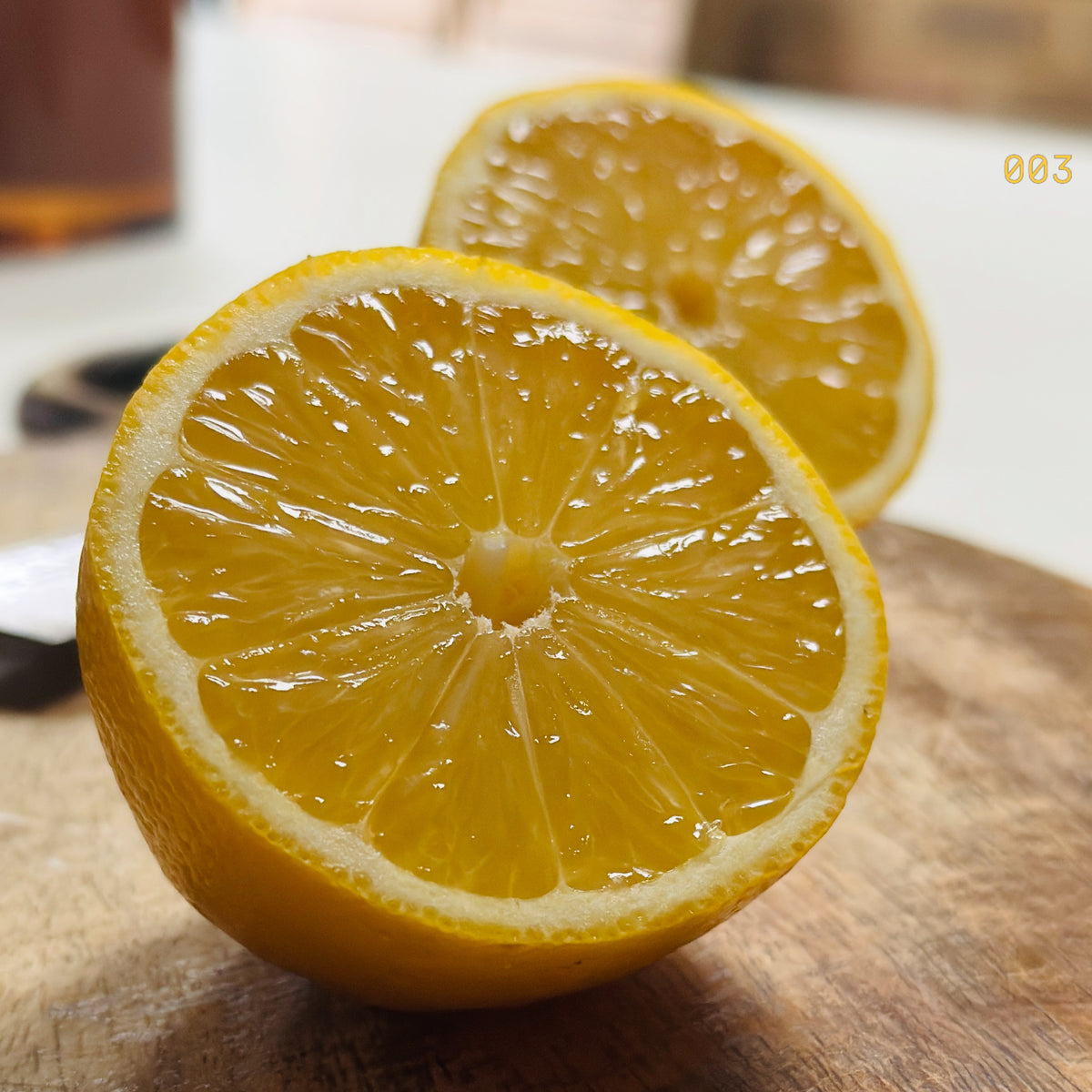 A close-up of a halved lemon on a wooden cutting board with a second lemon half blurred in the background. The lemon halves show bright yellow-orange pulp and rind, with a small yellow '003' text overlay in the top right corner.