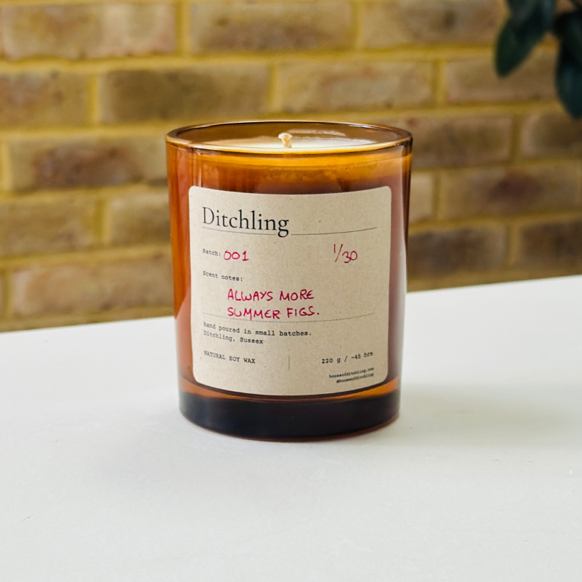 Ditchling “Always More Summer Figs” candle, Batch 001, 1/30, in an open amber glass jar showing the natural soy wax and wick. Label reads “Scent notes: ALWAYS MORE SUMMER FIGS.”, “hand poured in small batches. Ditchling, Sussex”, “NATURAL SOY WAX”, “220 g / ~45 hrs”, “houseofditchling.com” and “@houseofditchling”. Photographed on a white surface against a warm brick wall with greenery.