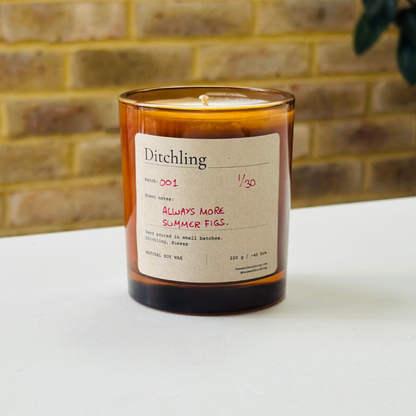 Ditchling “Always More Summer Figs” candle, Batch 001, 1/30, in an open amber glass jar showing the natural soy wax and wick. Label reads “Scent notes: ALWAYS MORE SUMMER FIGS.”, “hand poured in small batches. Ditchling, Sussex”, “NATURAL SOY WAX”, “220 g / ~45 hrs”, “houseofditchling.com” and “@houseofditchling”. Photographed on a white surface against a warm brick wall with greenery.