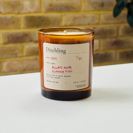 Ditchling “Always More Summer Figs” candle, Batch 001, 1/30, in an open amber glass jar showing the natural soy wax and wick. Label reads “Scent notes: ALWAYS MORE SUMMER FIGS.”, “hand poured in small batches. Ditchling, Sussex”, “NATURAL SOY WAX”, “220 g / ~45 hrs”, “houseofditchling.com” and “@houseofditchling”. Photographed on a white surface against a warm brick wall with greenery.