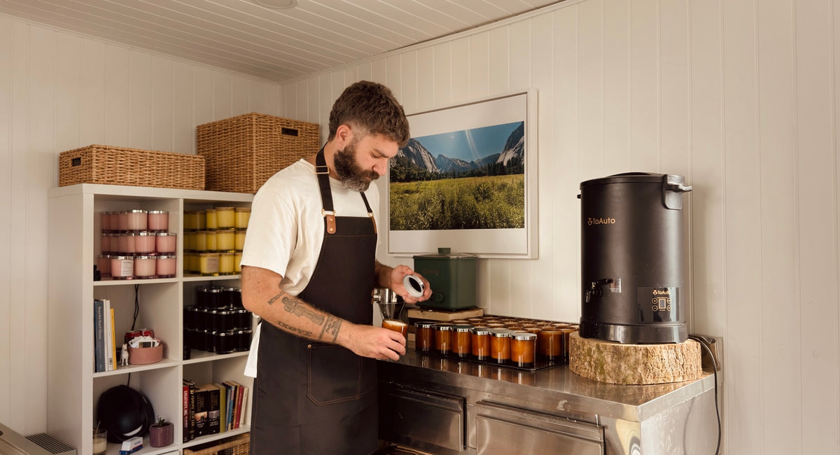 Person in a dark apron placing a lid on an amber candle jar at a stainless-steel candle-making station, with rows of amber jars, ToAuto wax melters, wicker baskets and a framed Yosemite landscape print in a bright white-panelled workshop.