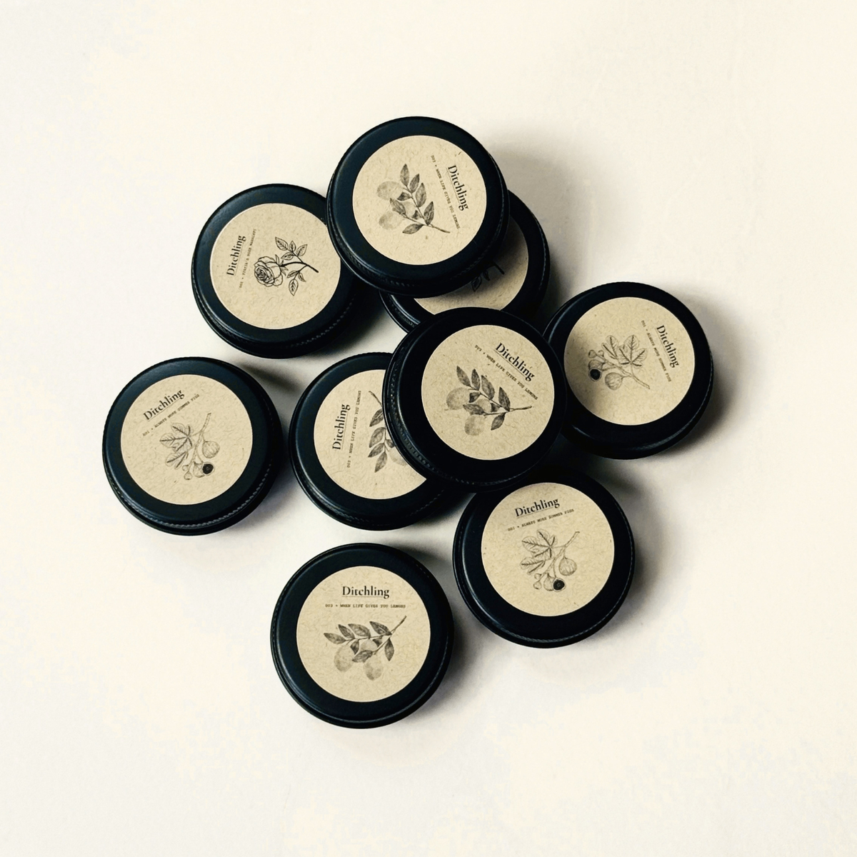 Ditchling 20ml sample candles in black tins, arranged together with kraft labels for 001 Always More Summer Figs, 002 Yiayia’s Rose Mahalepi and 003 When Life Gives You Lemons.