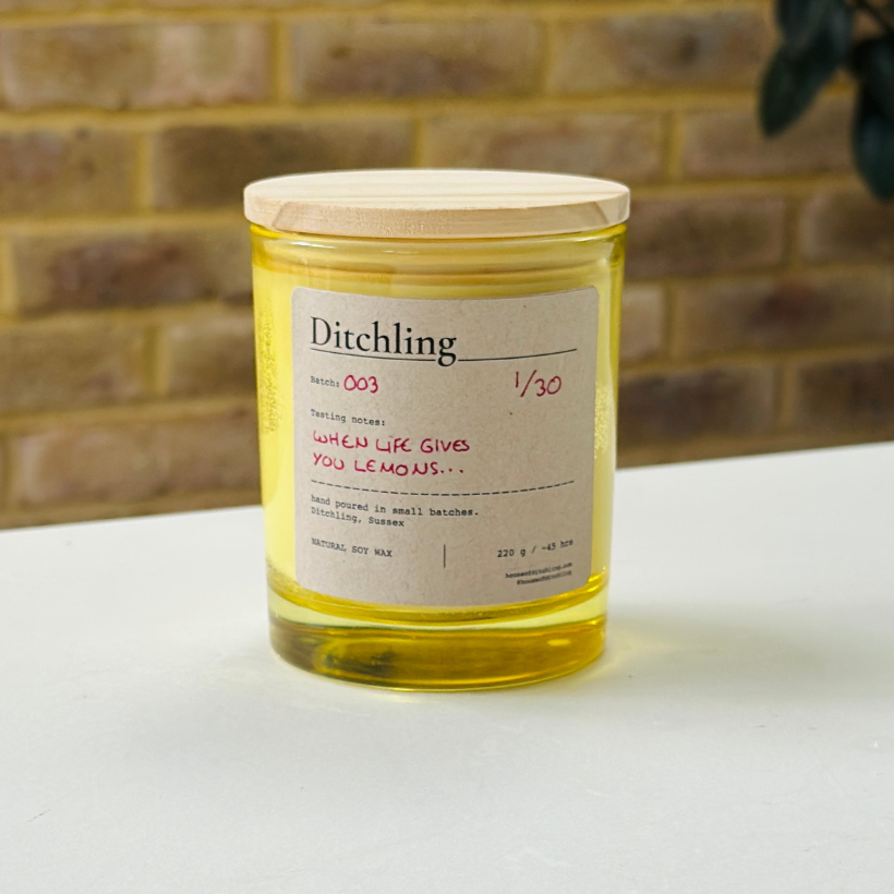 Ditchling “When Life Gives You Lemons…” candle, Batch 003, 1/30, in a yellow glass jar with wooden lid. Label reads “Tasting notes: WHEN LIFE GIVES YOU LEMONS…”, “hand poured in small batches. Ditchling, Sussex”, “NATURAL SOY WAX”, “220 g / ~45 hrs”, “houseofditchling.com” and “@houseofditchling”. Photographed on a white surface against a warm brick wall with greenery.