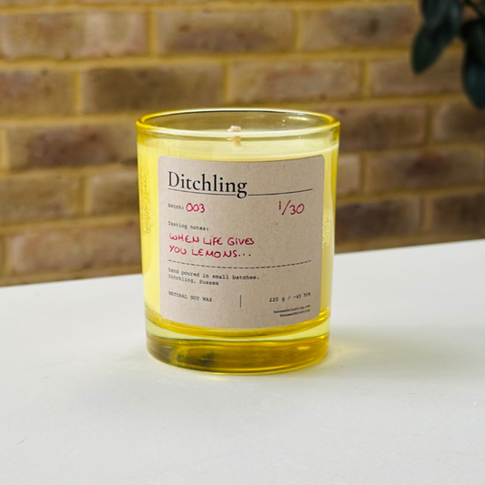 Ditchling “When Life Gives You Lemons…” candle, Batch 003, 1/30, in an open yellow glass jar showing the natural soy wax and wick. Label reads “Tasting notes: WHEN LIFE GIVES YOU LEMONS…”, “hand poured in small batches. Ditchling, Sussex”, “NATURAL SOY WAX”, “220 g / ~45 hrs”, “houseofditchling.com” and “@houseofditchling”. Photographed on a white surface against a warm brick wall with greenery.