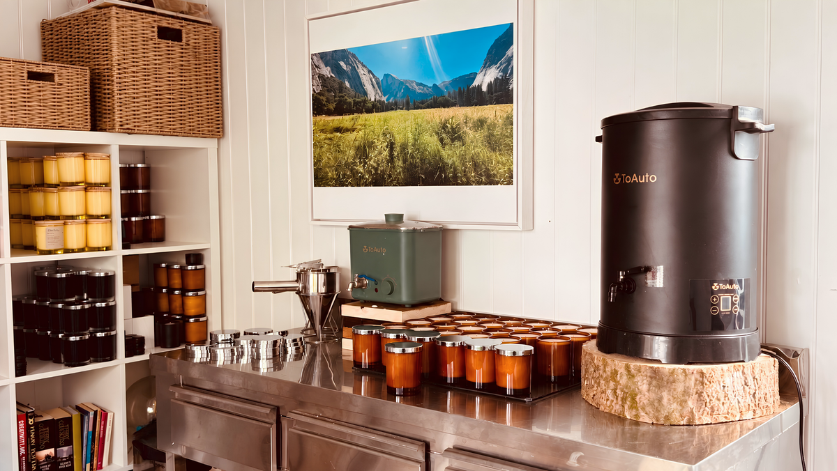 Candle-making workshop with rows of amber glass candles on a stainless-steel workbench, ToAuto wax melters, metal pouring tools, rattan storage baskets and a framed Yosemite landscape photograph on the wall.