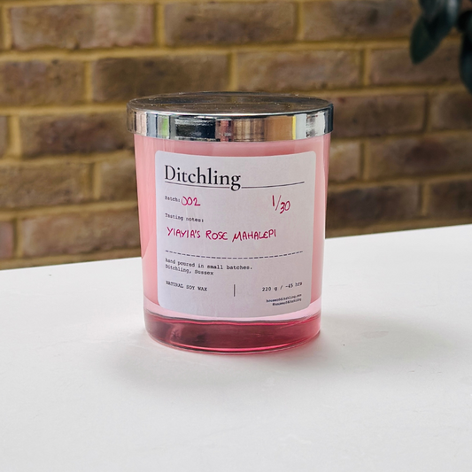 Ditchling “Yiayia’s Rose Mahalepi” candle, Batch 002, 1/30, in a pink glass jar with silver lid. Label reads “Tasting notes: YIAYIA’S ROSE MAHALEPI”, “hand poured in small batches. Ditchling, Sussex”, “NATURAL SOY WAX”, “220 g / ~45 hrs”, “houseofditchling.com” and “@houseofditchling”. Photographed on a white surface against a warm brick wall with greenery.