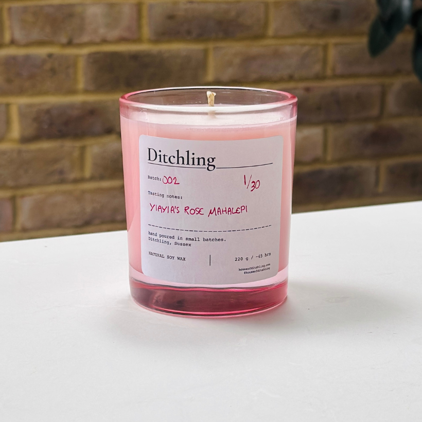 Ditchling “Yiayia’s Rose Mahalepi” candle, Batch 002, 1/30, in an open pink glass jar showing the natural soy wax and wick. Label reads “Tasting notes: YIAYIA’S ROSE MAHALEPI”, “hand poured in small batches. Ditchling, Sussex”, “NATURAL SOY WAX”, “220 g / ~45 hrs”, “houseofditchling.com” and “@houseofditchling”. Photographed on a white surface against a warm brick wall with greenery.